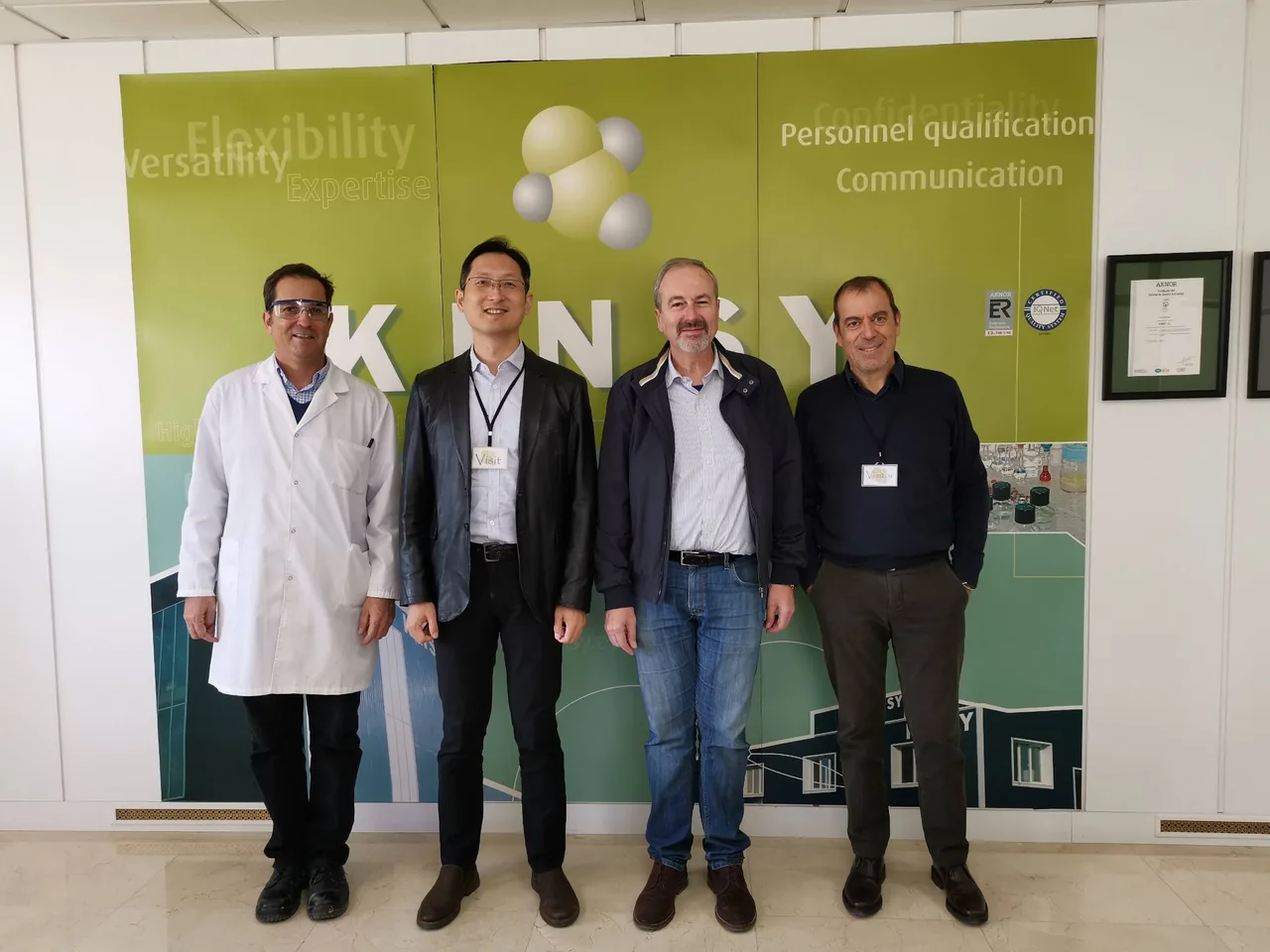 A visit to our customer KINRAY's plant: four colleagues pose in front of the KINRAY brand wall, one in a lab coat