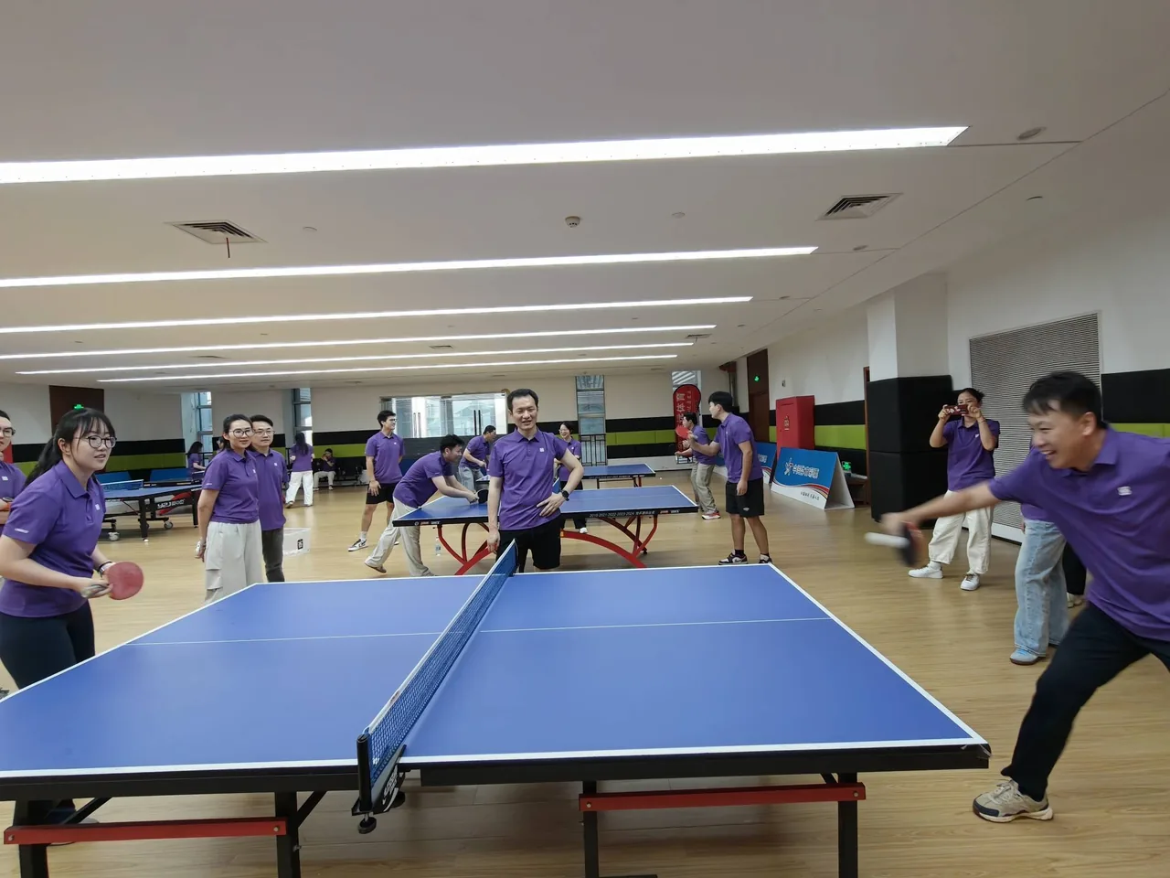Several tables in play inside the table-tennis hall; in the foreground two colleagues rally while teammates in the brand purple kit watch on, one filming with a phone